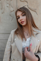 Fashion beautiful girl with a Caucasian face in fashionable clothes with a stylish leather jacket and a white top stands near a vintage column in the park