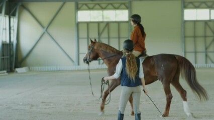 Woman riding chestnut horse lead by female instructor during lesson in equestrian school