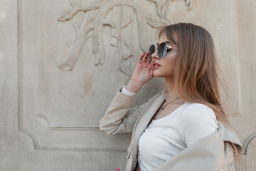 Fashionable female portrait of a beautiful young stylish girl in fashion casual outfit with leather jacket and top wearing cool sunglasses near a vintage wall