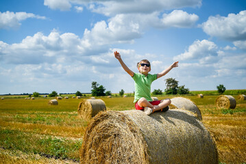 Naklejka na ściany i meble Little boy sits on a round haystack relaxed with hands up. Field