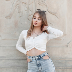 Stylish cute beauty young woman hipster in summer fashion clothes with top and jeans stands near a vintage wall outdoors
