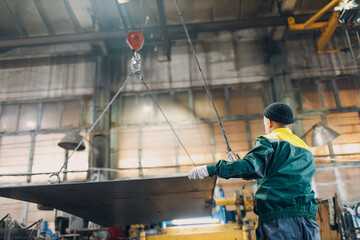 Worker lift metal sheet with crane chain hoist with remote controller and hook at industrial...