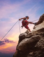 Foto op Plexiglas Muscular climber man in protective helmet abseiling from cliff rock wall using rope Belay device and climbing harness on evening sunset sky background. Active extreme sports time spending concept. © Soloviova Liudmyla