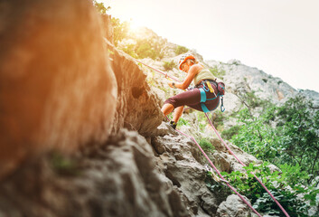 Active climber woman in protective helmet abseiling from cliff rock wall using rope with belay...
