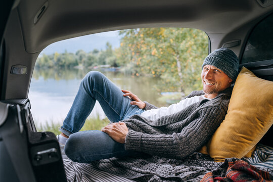 Midle-aged smiling man dressed in warm knitted clothes lying in opened Car trunk and enjoying a beautiful autumnal mountain lake view. Warm early autumn auto traveling concept image.
