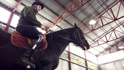 in special hangar, a young disabled man learns to ride a black, thoroughbred horse, hippotherapy. man has an artificial limb instead of his right leg. concept of rehabilitation of disabled with - obrazy, fototapety, plakaty
