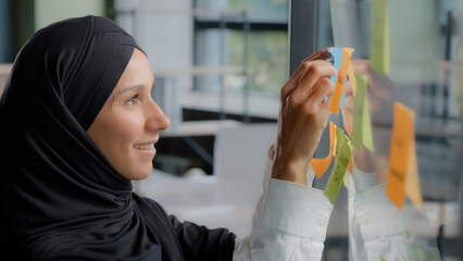 Smiling successful businesswoman corporate leader writing project tasks leaves reminder on sticky colored note organizes work young arab woman office worker in hijab attaches stickers on glass board