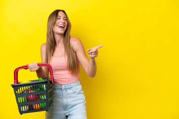 Young caucasian woman holding a shopping basket full of food isolated on yellow background pointing finger to the side and presenting a product