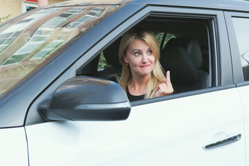 Young woman driving a car stuck her head out the window and showing middle finger. Angry woman with blond hair demonstrating fuck you off sign from open window.