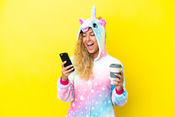 Girl with curly hair wearing a unicorn pajama isolated on yellow background holding coffee to take away and a mobile © luismolinero