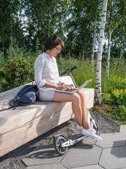 Fototapeta premium Mindful woman works with laptop on wooden bench in urban park. Summer vibes. Modern workplace for freelancers. Student educates online from outdoors.