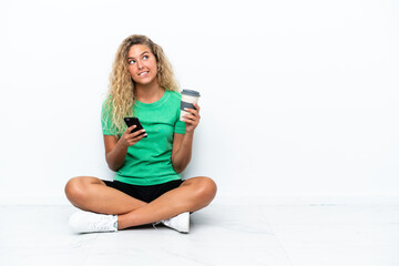 Girl with curly hair sitting on the floor holding coffee to take away and a mobile while thinking something