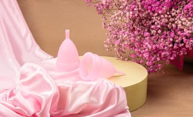 Two pink menstrual cups made of medical silicone lie on a cylindrical podium, surrounded by delicate pink satin, on a beige craft paper background.  Small flowers nearby