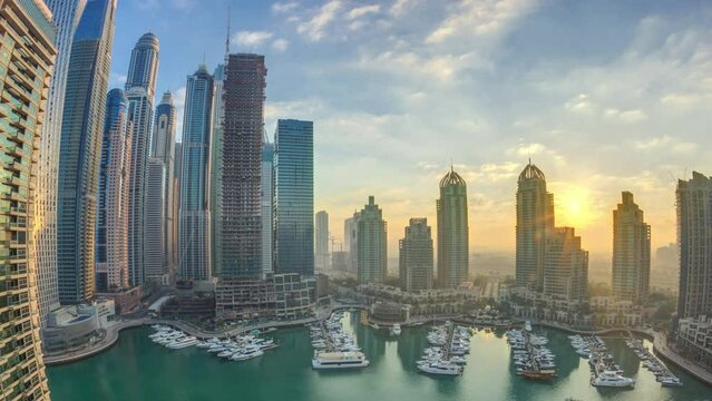 Panorama of modern skyscrapers shining in sunrise lights timelapse in Dubai Marina with yachts in Dubai, UAE. Rays of lights behind buildings. Orange clouds on the sky