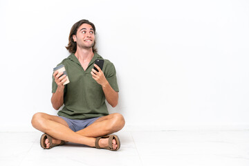 Young handsome man sitting on the floor isolated on white background holding coffee to take away and a mobile while thinking something
