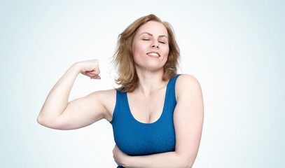 Fototapeta premium Young woman in a tight blue dress shows the muscles on her arm, smiling broadly and closing her eyes, isolated on light blue background
