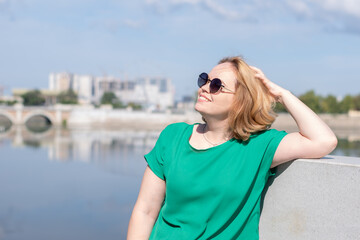 Portrait of a girl in sunglasses, a green blouse standing on the riverbank holding her hair with her hand.