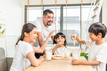 Fototapeta premium Asian family enjoying breakfast at living room. little girl daughter sitting on table, drinking milk with smiling father and mother in morning. Happy family at home.