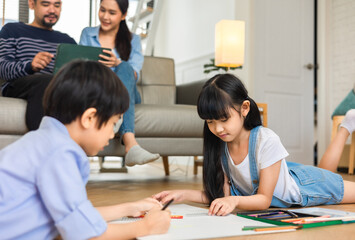 Happy Asian family relaxing on sofa while kid drawing on floor. Little boy girl having fun, friendship between siblings, family leisure time in living room at home.