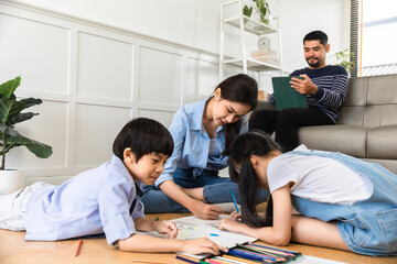 Happy Asian family relaxing on sofa while kid drawing on floor. Little boy girl having fun, friendship between siblings, family leisure time in living room at home.