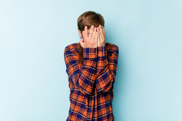 Young caucasian red-haired man isolated on blue background blink at the camera through fingers, embarrassed covering face.