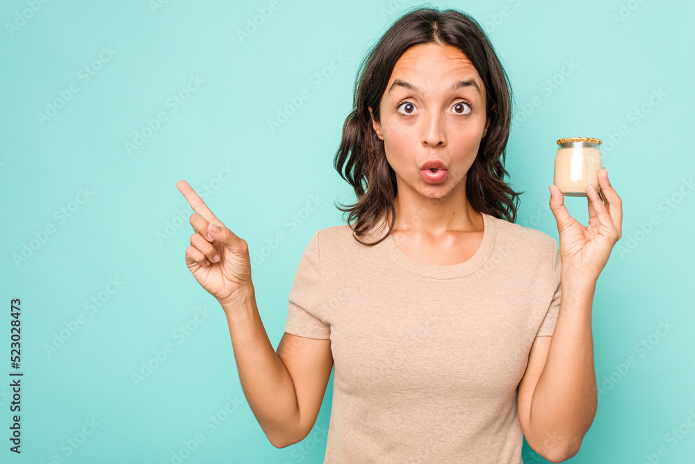 Wall mural Young hispanic woman holding yogurt isolated on blue background pointing to the side