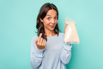 Young hispanic woman holding a bag of oats isolated on blue background pointing with finger at you as if inviting come closer.