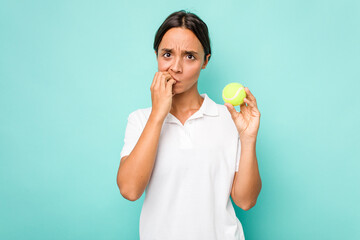 Young hispanic physiotherapy holding a tennis ball isolated on blue background biting fingernails, nervous and very anxious.