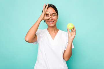 Young hispanic physiotherapy holding a tennis ball isolated on blue background excited keeping ok gesture on eye.