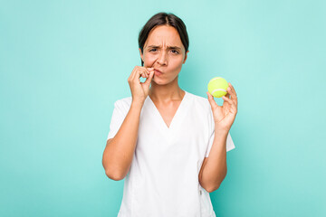 Young hispanic physiotherapy holding a tennis ball isolated on blue background with fingers on lips keeping a secret.