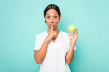 Young hispanic physiotherapy holding a tennis ball isolated on blue background keeping a secret or asking for silence.