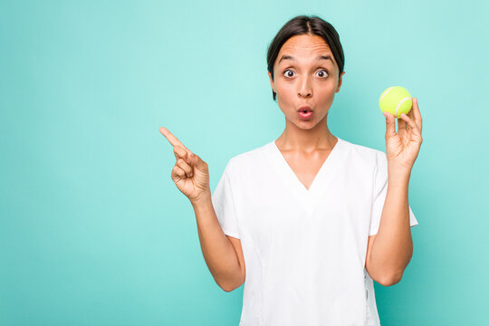 Young hispanic physiotherapy holding a tennis ball isolated on blue background pointing to the side