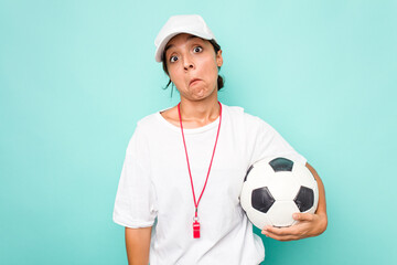Young hispanic soccer referee woman isolated on blue background shrugs shoulders and open eyes...