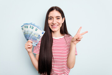 Young caucasian woman holding banknotes isolated on blue background joyful and carefree showing a peace symbol with fingers.