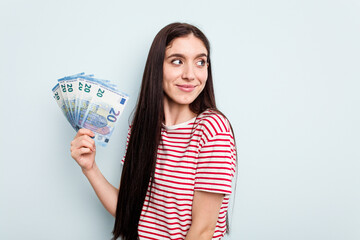 Young caucasian woman holding banknotes isolated on blue background looks aside smiling, cheerful and pleasant.