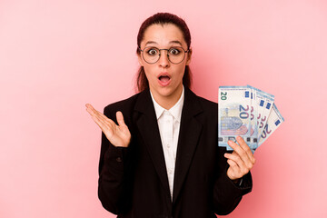 Young business caucasian woman holding bank notes isolated on pink background surprised and shocked.