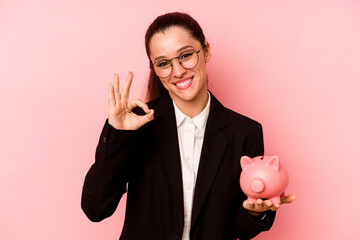 Young business caucasian woman holding piggybank isolated on pink background cheerful and confident...