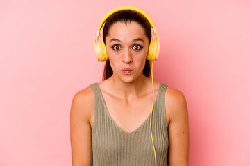 Young caucasian woman listening to music isolated on pink background shrugs shoulders and open eyes...