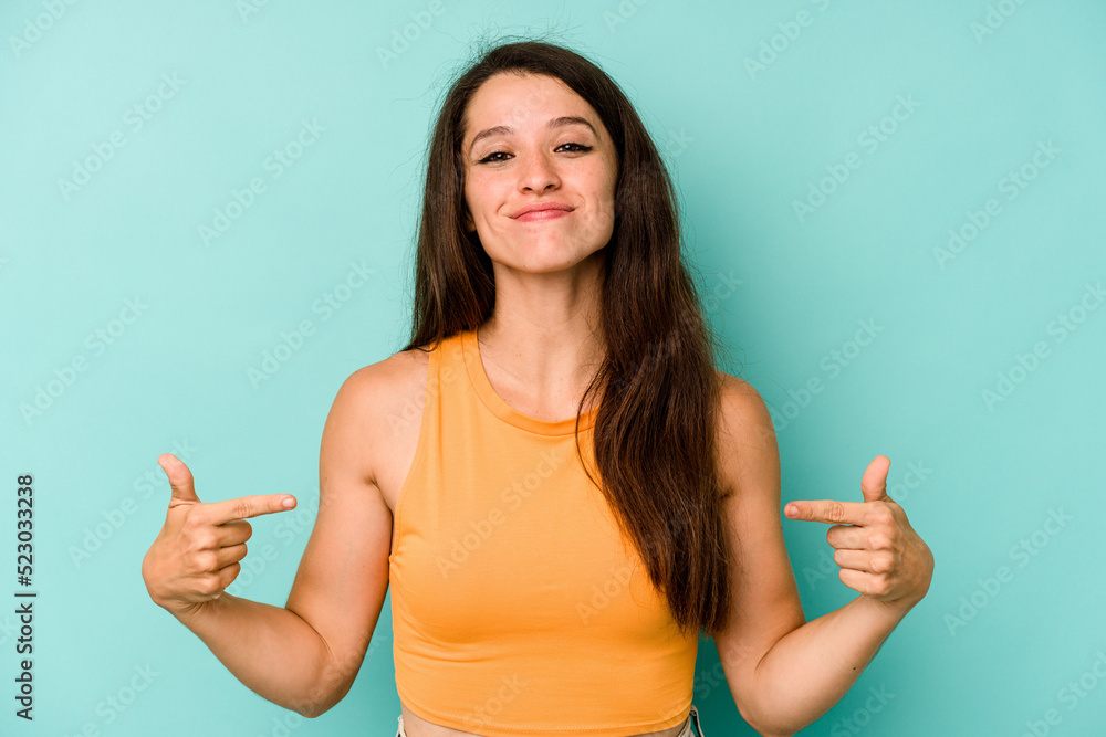 Wall mural Young caucasian woman isolated on blue background person pointing by hand to a shirt copy space, proud and confident