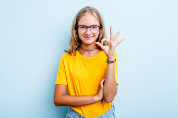 Caucasian teen girl isolated on blue background winks an eye and holds an okay gesture with hand.