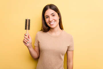 Young caucasian woman holding a brush hair isolated on yellow background happy, smiling and cheerful.