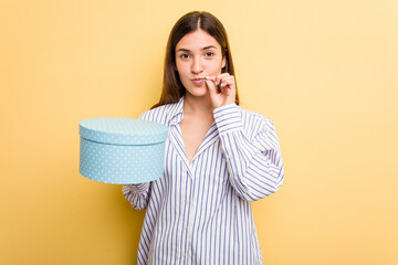 Young caucasian woman holding a gift box isolated on yellow background with fingers on lips keeping a secret.