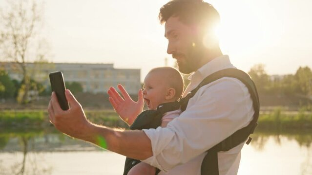 At sunlight young father with a little newborn baby in a slings carrier. Use phone to take selfies on street near lake in park. Ergo backpack. Parents. Gadget. Smartphone. Cell. Slow motion