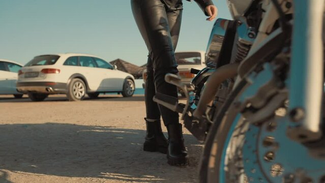 Woman biker in leather pants approaches the motorcycle and sits on it. Slow motion. Low angle view. Truck shot. The concept of motorcycle trips and freedom.