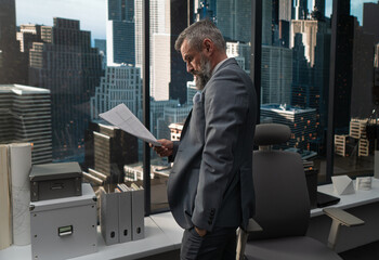 Portrait of 50s adult grey-haired Caucasian CEO businessman looking through papers near window in the office located in a skyscraper, business district buildings in the background