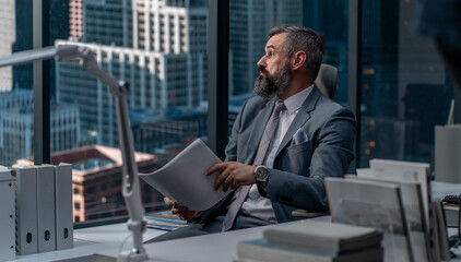 Portrait of 50s adult grey-haired Caucasian CEO businessman working at his desk in the office located in a skyscraper, business district buildings in the background