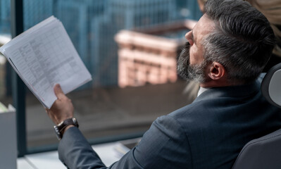 Portrait of 50s adult grey-haired Caucasian CEO businessman working at his desk in the office located in a skyscraper, business district buildings in the background