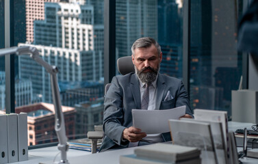 Fototapeta premium Portrait of 50s adult grey-haired Caucasian CEO businessman working at his desk in the office located in a skyscraper, business district buildings in the background