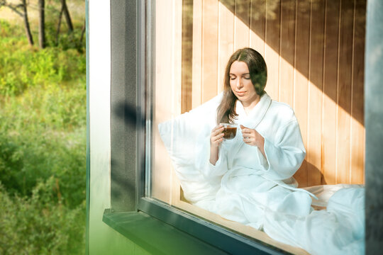 Nature retreat, comfortable unity with nature. Young woman sitting on the window of beautiful country house or eco house, holding cup of coffee. Outside view through the glass window.