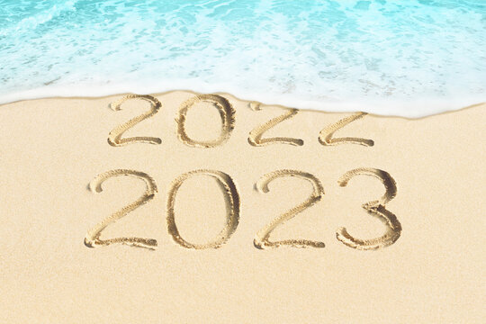 New Year concept photo of coming 2023 and leaving 2022. Blue sea wave washing away the handwritten in sand numbers. Sandy beach background.
